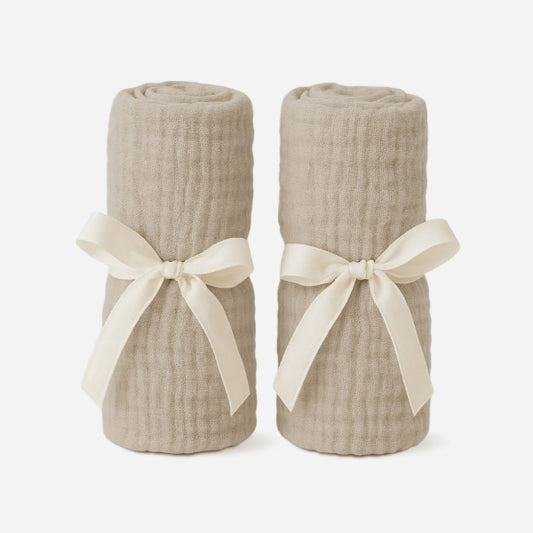 Two beige, rolled-up blankets stand upright, each tied with a cream ribbon, perfectly complementing your Newton Baby Compact Travel Crib Sheets—shown against a plain white background.