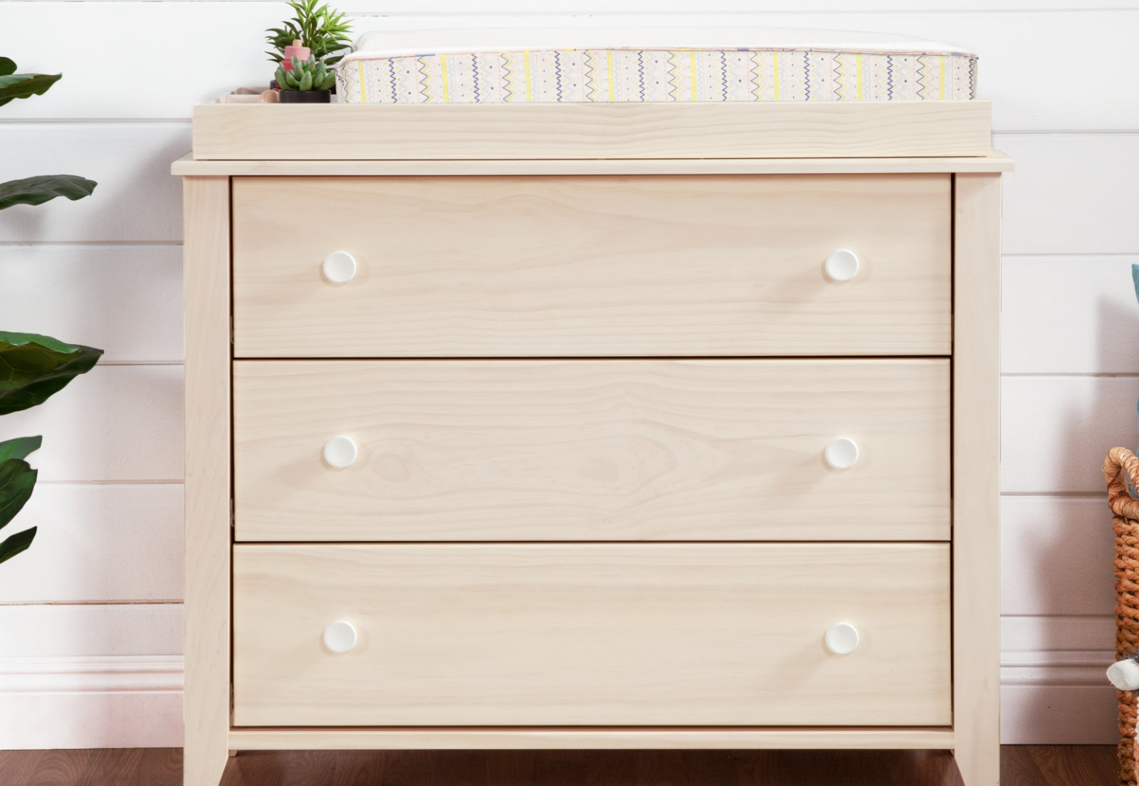 A light wood changing table dresser with three drawers and white knobs, featuring a small plant and a striped changing pad on top, placed in a nursery room. 999-8603-WWH