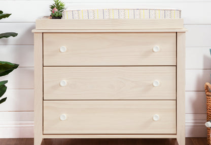 A light wood changing table dresser with three drawers and white knobs, featuring a small plant and a striped changing pad on top, placed in a nursery room. 999-8603-WWH