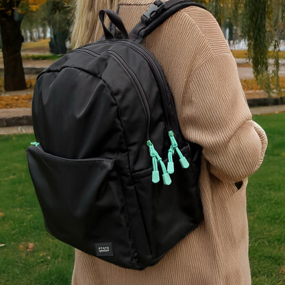 A person in a beige coat carries the Newton Lorimer Diaper Bag by Newton Baby, featuring mint green zipper pulls, while standing on grass with trees and autumn leaves in the background.