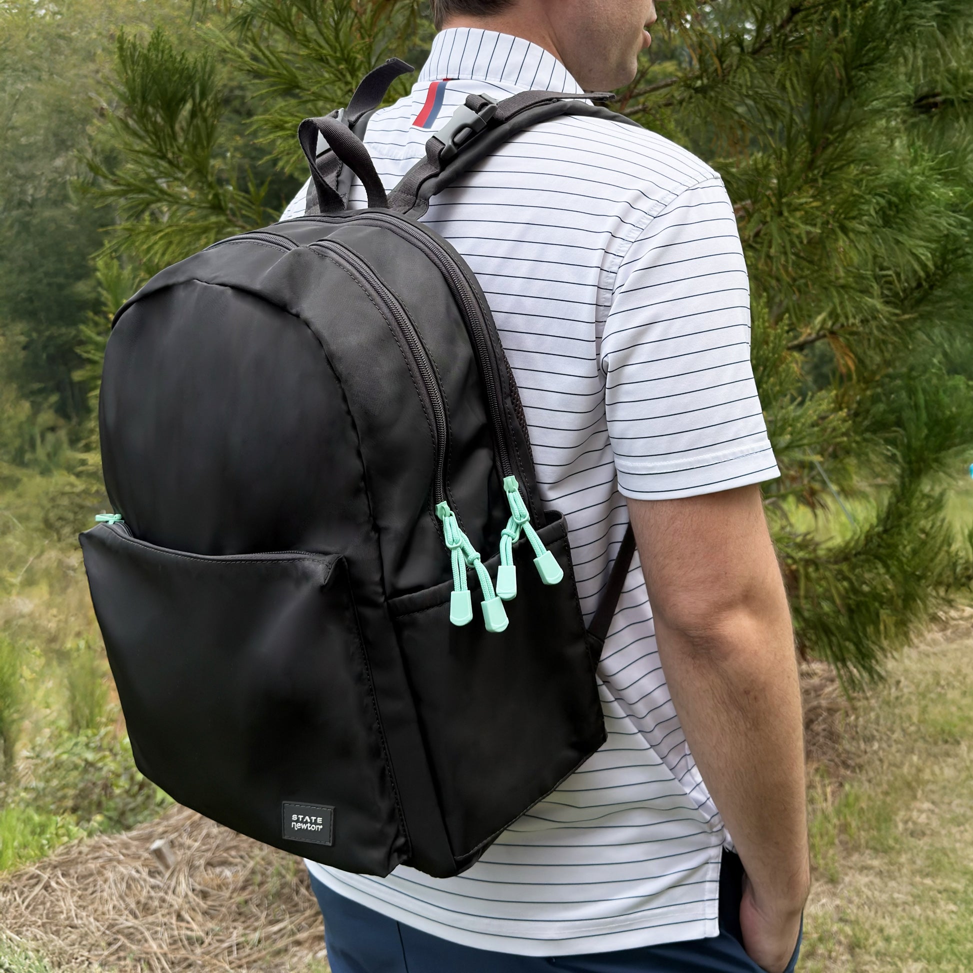 Outdoors among trees, a person wears a white striped short-sleeve shirt and carries the spacious Newton Lorimer Diaper Bag by STATE from Newton Baby, featuring light blue zipper pulls.