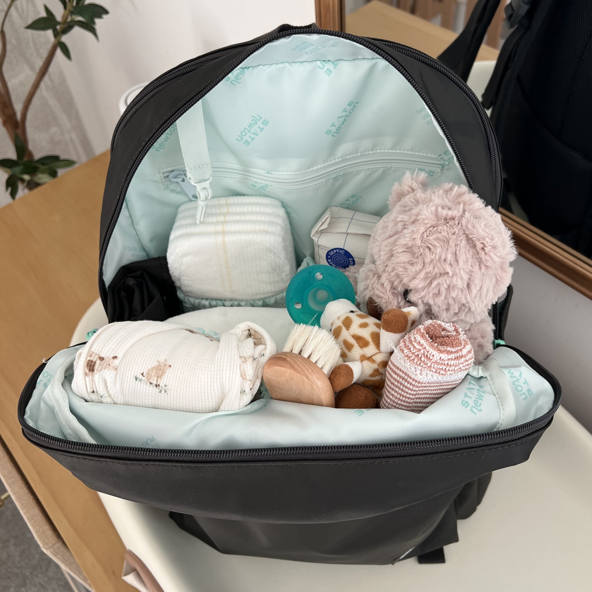 The Newton Lorimer Diaper Bag by STATE from Newton Baby sits open on a table, filled with baby must-haves like a plush bear, diapers, pacifier, bottle, teether, swaddle, and striped socks, with a plant and mirror in the background.