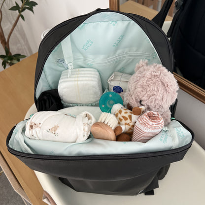 The Newton Lorimer Diaper Bag by STATE from Newton Baby sits open on a table, filled with baby must-haves like a plush bear, diapers, pacifier, bottle, teether, swaddle, and striped socks, with a plant and mirror in the background.
