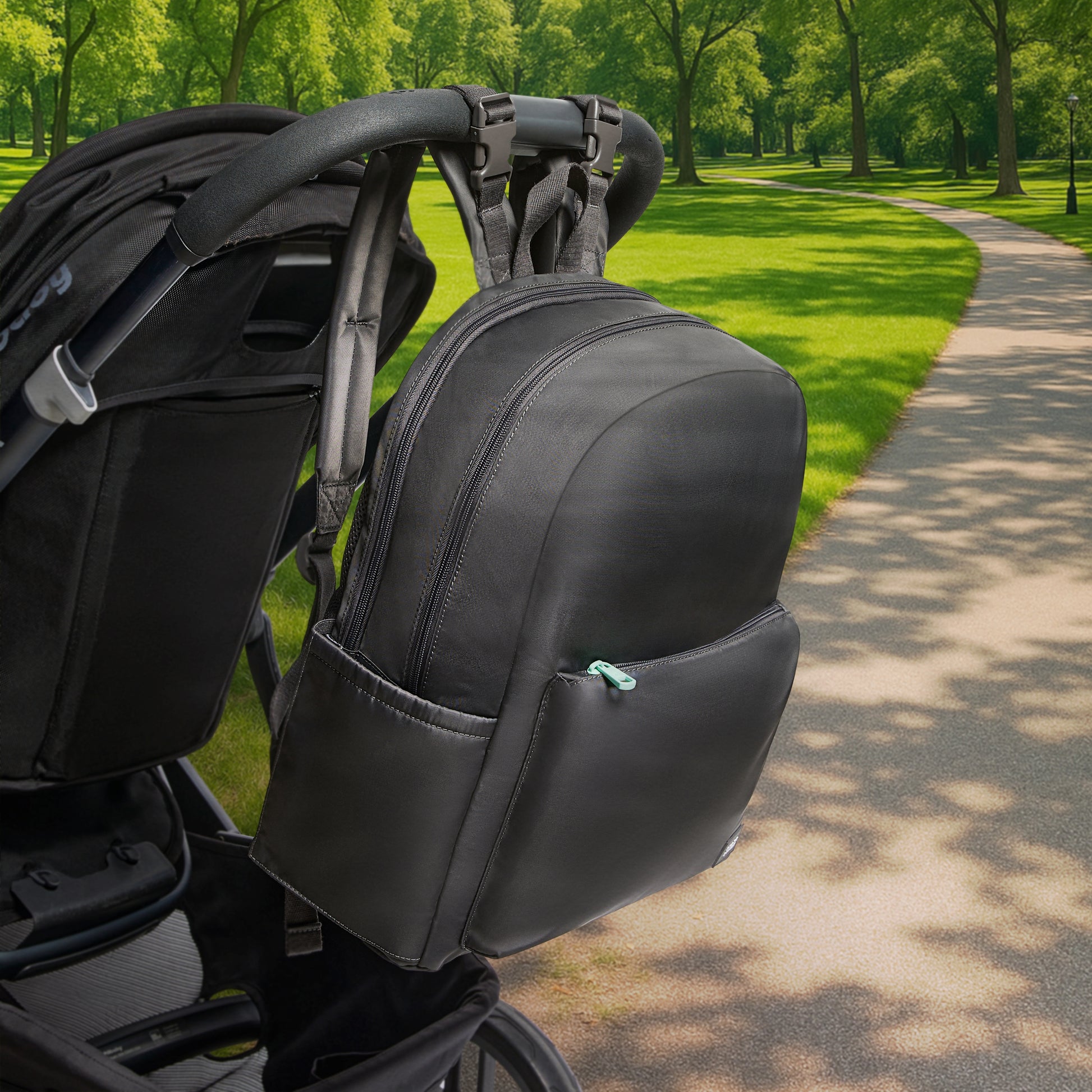 The Newton Lorimer Diaper Bag by Newton Baby hangs from a stroller handlebar on a paved path in a sunny, green park.