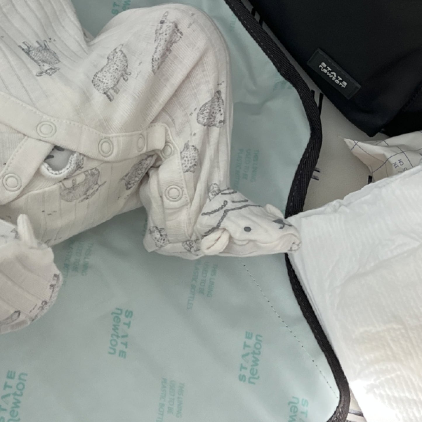 A baby in white animal-print pajamas lies on a changing mat, with only legs and feet visible. Nearby are a diaper and the Newton Lorimer Diaper Bag by STATE from Newton Baby, ready for any quick change or need.