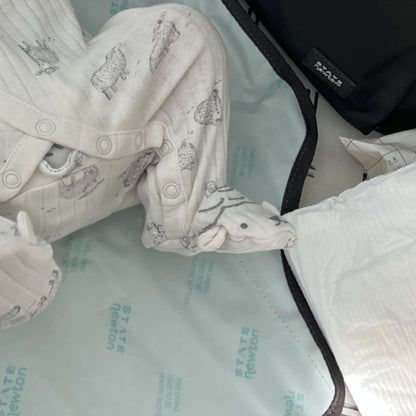A baby in white animal-print pajamas lies on a changing mat, with only legs and feet visible. Nearby are a diaper and the Newton Lorimer Diaper Bag by STATE from Newton Baby, ready for any quick change or need.