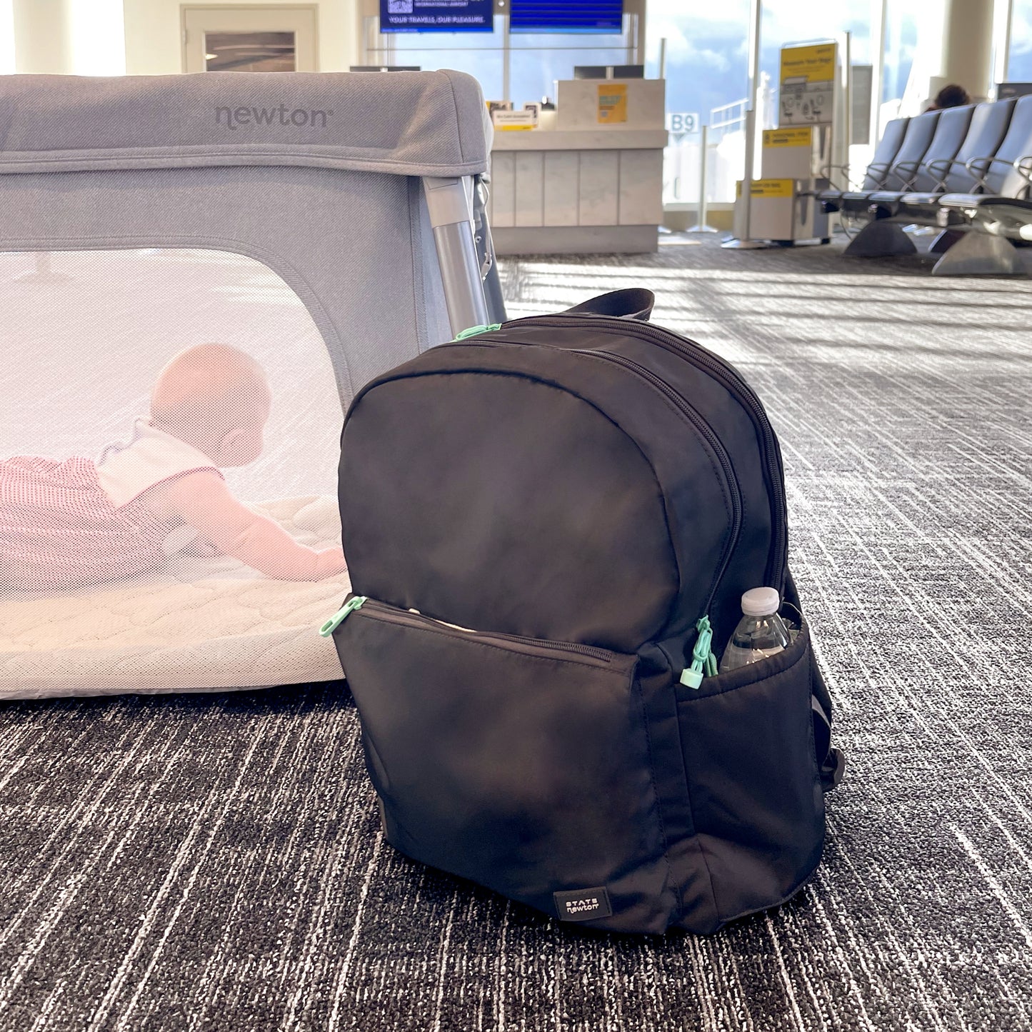 A black Newton Lorimer Diaper Bag by Newton Baby sits on the airport floor with a water bottle in its side pocket. In the background, a baby crawls inside a gray playpen near seats and a boarding gate.
