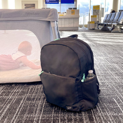 A black Newton Lorimer Diaper Bag by Newton Baby sits on the airport floor with a water bottle in its side pocket. In the background, a baby crawls inside a gray playpen near seats and a boarding gate.