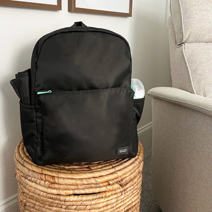 The Newton Lorimer Diaper Bag by Newton Baby sits on a woven basket by a light armchair, with a baby bottle visible in one of the spacious side pockets.