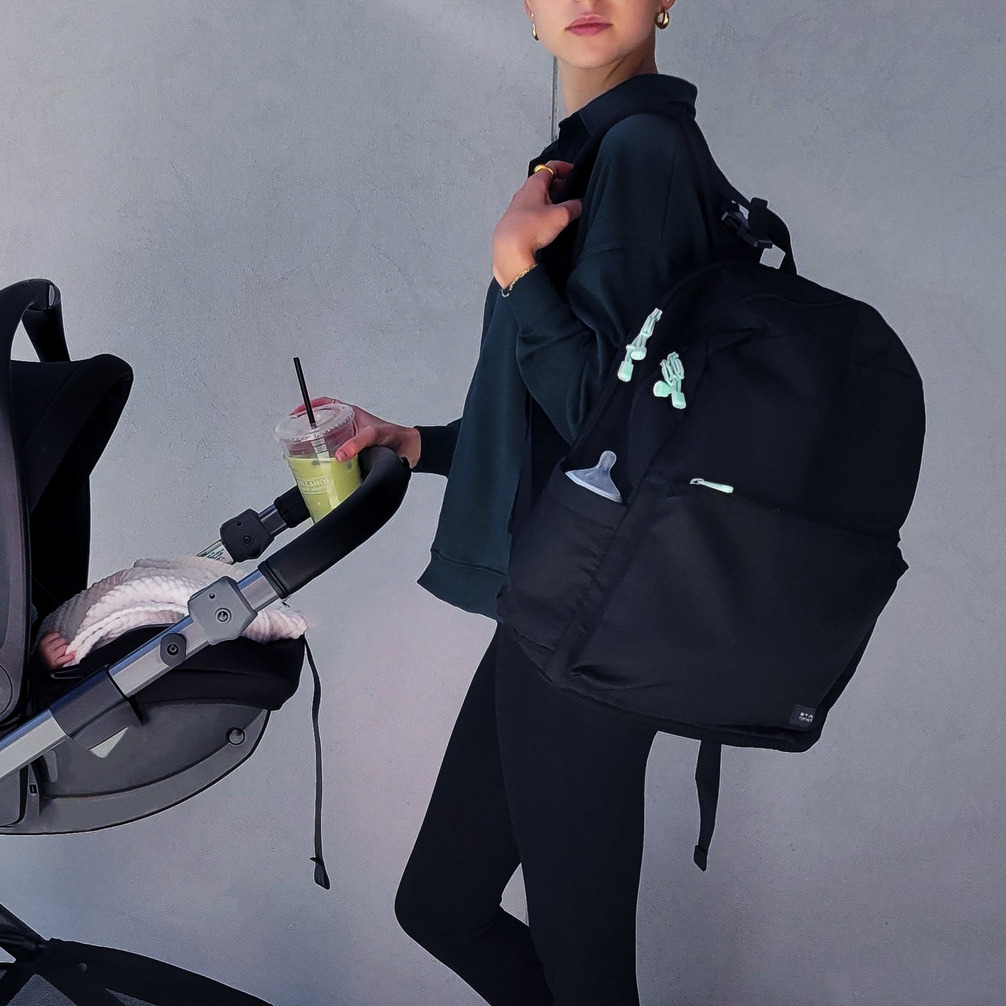 A woman in dark clothing carries a Newton Baby X STATE Lorimer Diaper Bag and a green drink while pushing a stroller with a pink blanket inside, against a light gray wall.