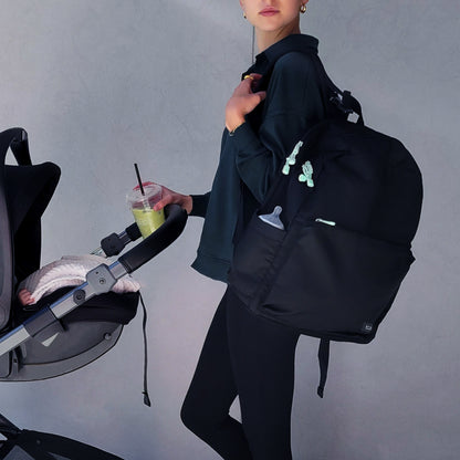 A woman in dark clothing carries a Newton Baby X STATE Lorimer Diaper Bag and a green drink while pushing a stroller with a pink blanket inside, against a light gray wall.