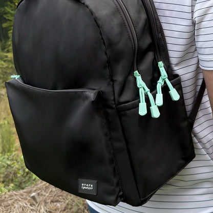 A person stands outdoors wearing a black Newton X STATE Lorimer Diaper Bag by Newton Baby, featuring light green zipper pulls, two large compartments, a small front pocket, and a breathable mesh back panel for comfort.