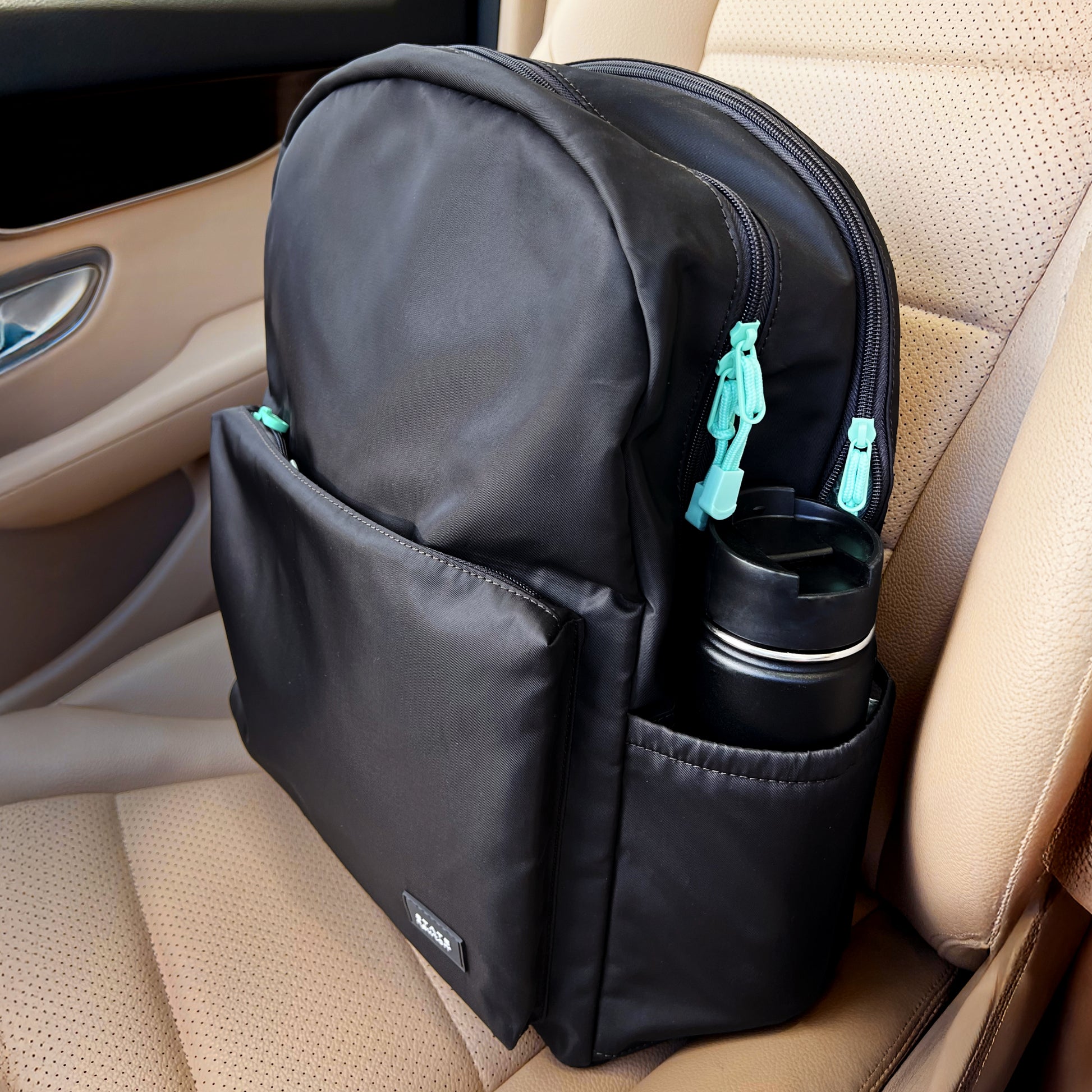 A stylish Newton Baby Newton X STATE Lorimer Diaper Bag with turquoise zippers rests on a tan car seat. It features an outer pocket, breathable mesh back panel, and a stainless steel travel mug in the side pocket—perfect for parents on the go.
