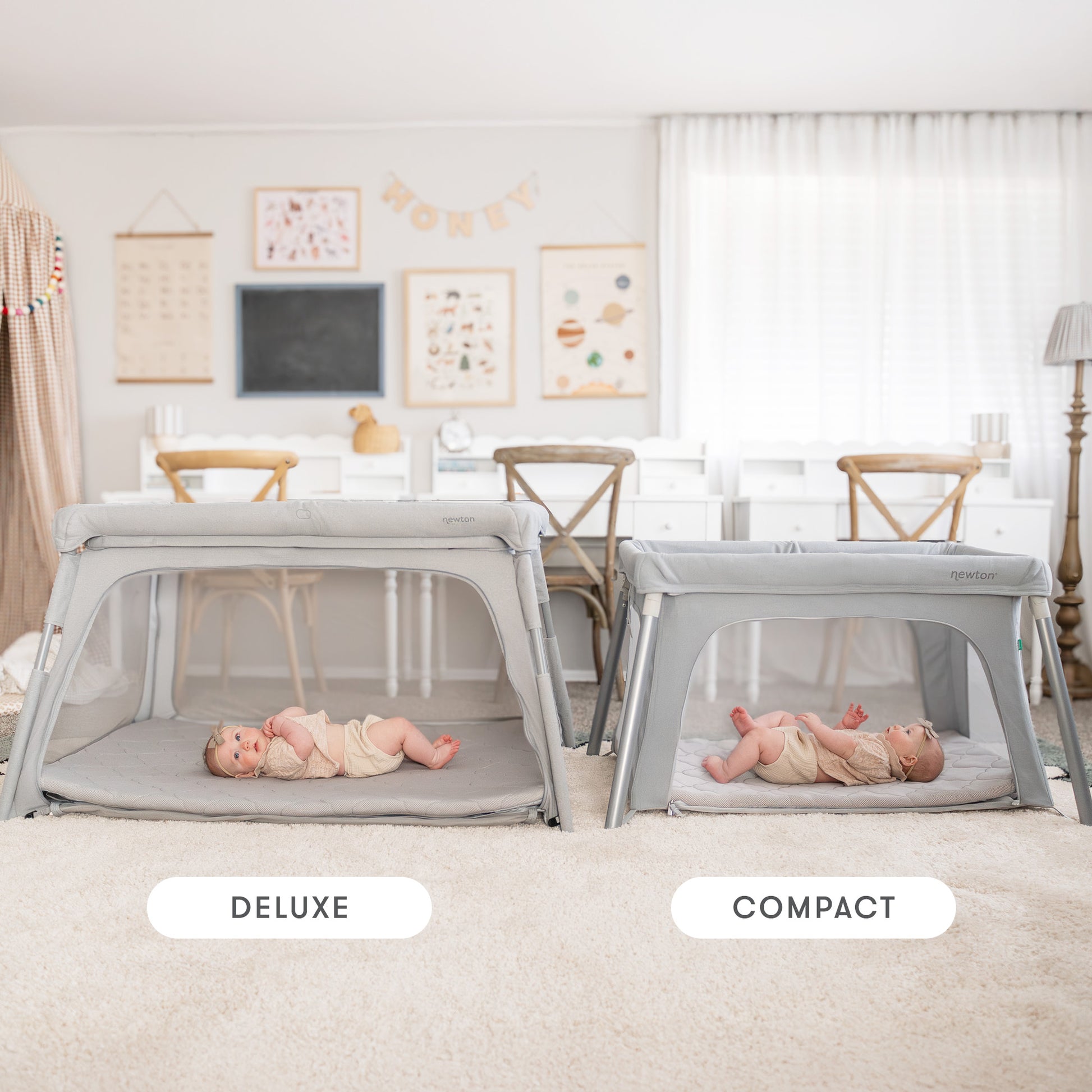 Two babies relax in Deluxe Travel Crib & Play Yards by Newton Baby, each with a breathable mattress, in a bright room with children's art, wooden chairs, and white curtains that let in soft natural light.