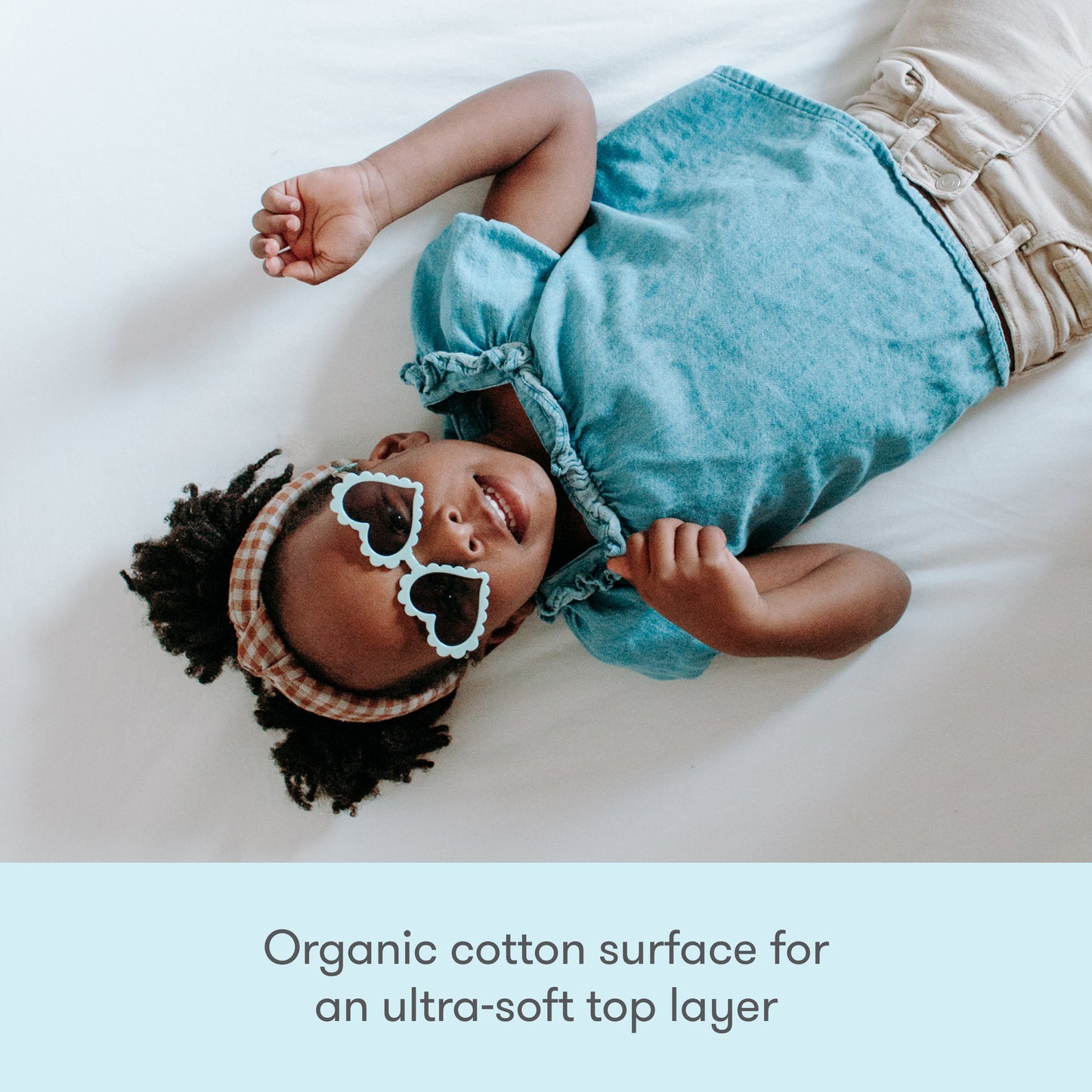 A young child with heart-shaped sunglasses and headband smiles on a white bed. Text: Newton Baby Waterproof Organic Twin Mattress Pad—ultra-soft, washable, waterproof protection for kids' beds.