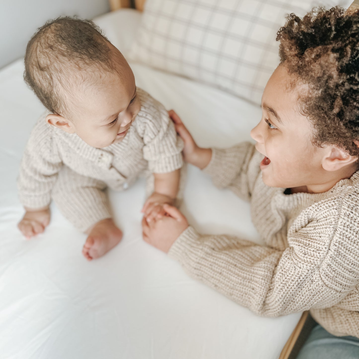 Two young children in matching beige knits sit on a bed, smiling and holding hands atop the Newton Baby Waterproof Organic Twin Mattress Pad, creating a cozy and joyful atmosphere.
