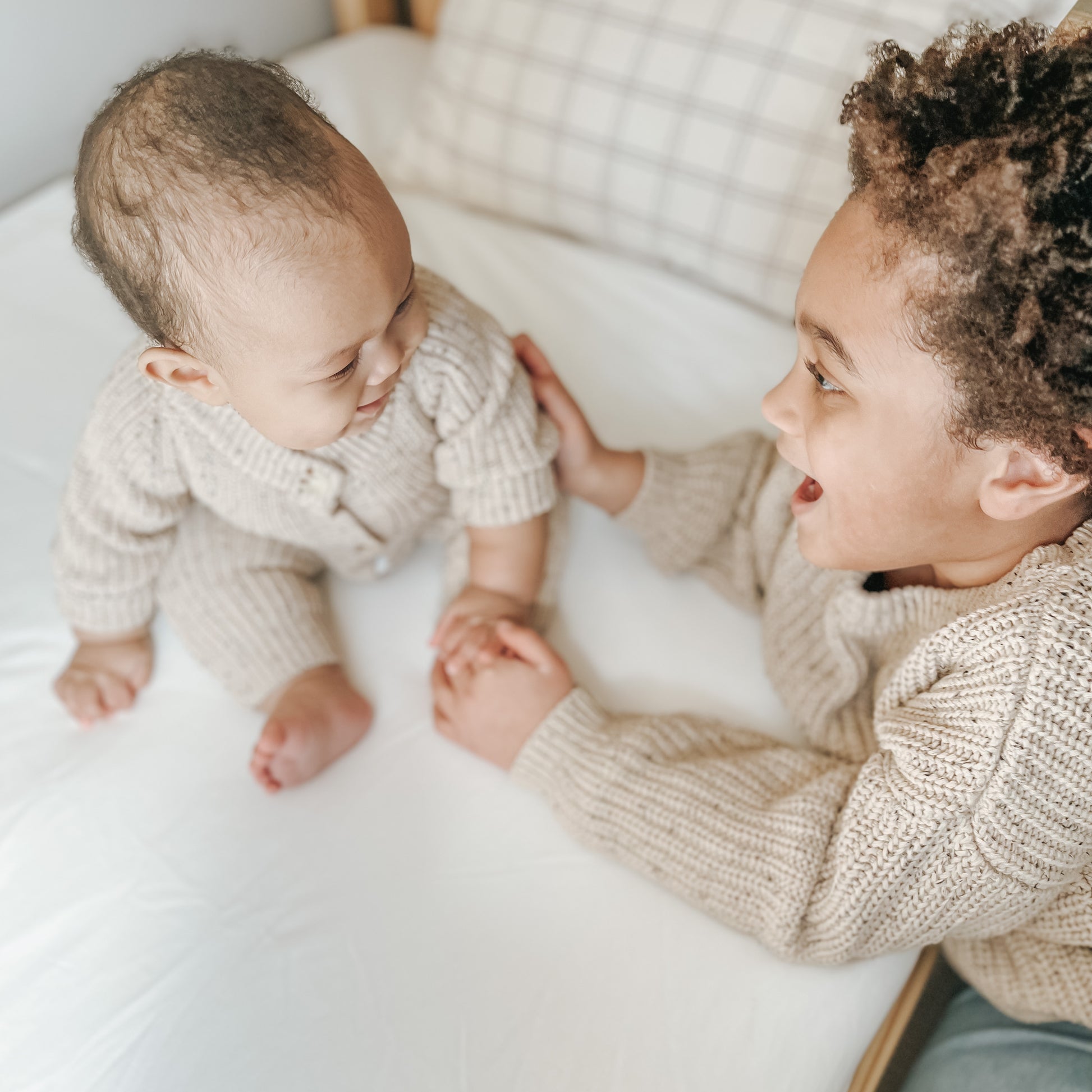 Two young children in matching beige knits sit on a bed, smiling and holding hands atop the Newton Baby Waterproof Organic Twin Mattress Pad, creating a cozy and joyful atmosphere.