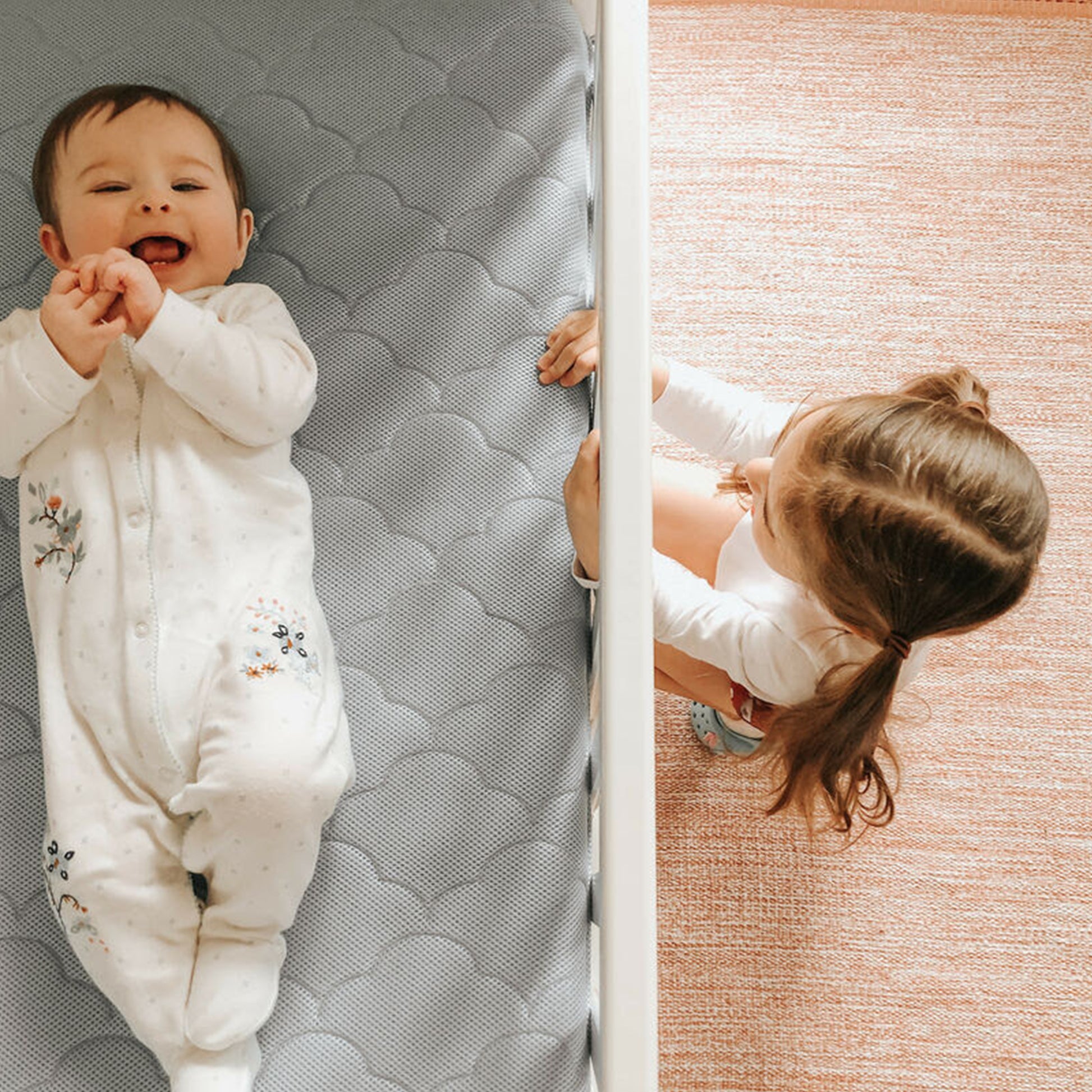 baby lying on back on mattress laughing with sister peering through crib at baby