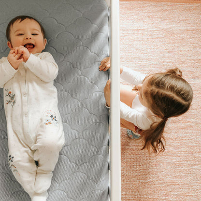 baby lying on back on mattress laughing with sister peering through crib at baby