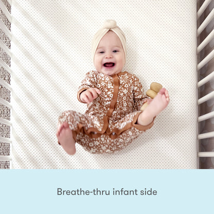 A smiling baby in a floral onesie and cream headband rests on the Newton Baby Essential Waterproof Crib Mattress. Text below reads, "Breathe-thru infant side.