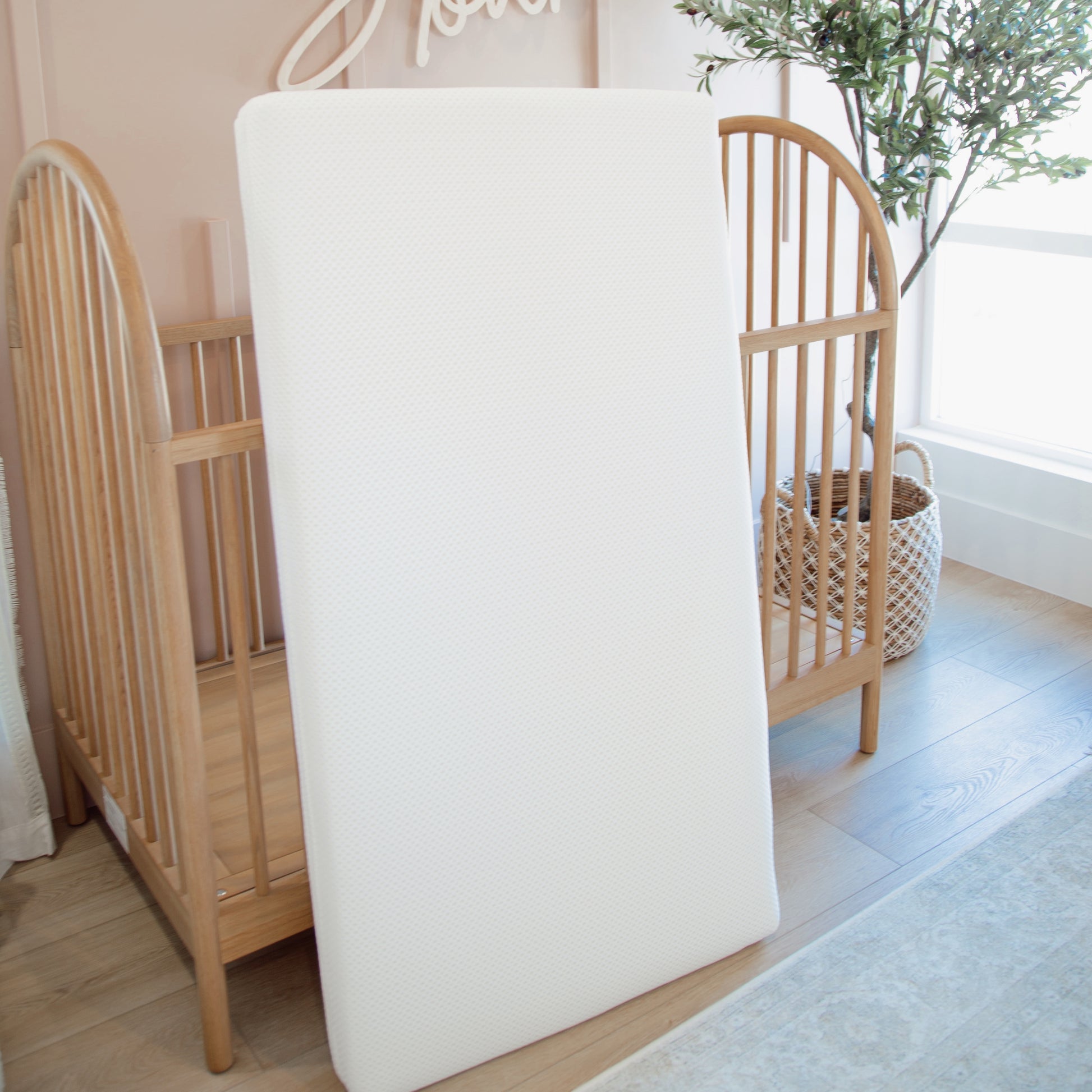 The Newton Baby Essential Waterproof Crib Mattress stands upright against a wooden crib in a bright, softly lit nursery, with a potted plant and window nearby.