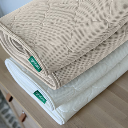 Two rolled-up Newton Baby Waterproof Crib Mattresses, one beige and one white, with quilted mesh, washable covers, and green Newton tags are stacked on a wooden surface.