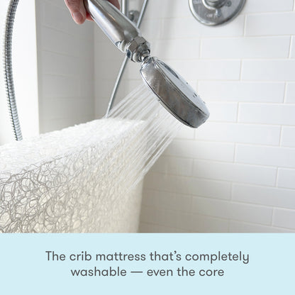 A handheld showerhead sprays water onto a Newton Baby Waterproof Crib Mattress core in a tiled bathroom, demonstrating this mattress is fully washable. Text below reads: The waterproof crib mattress that’s completely washable—even the core.