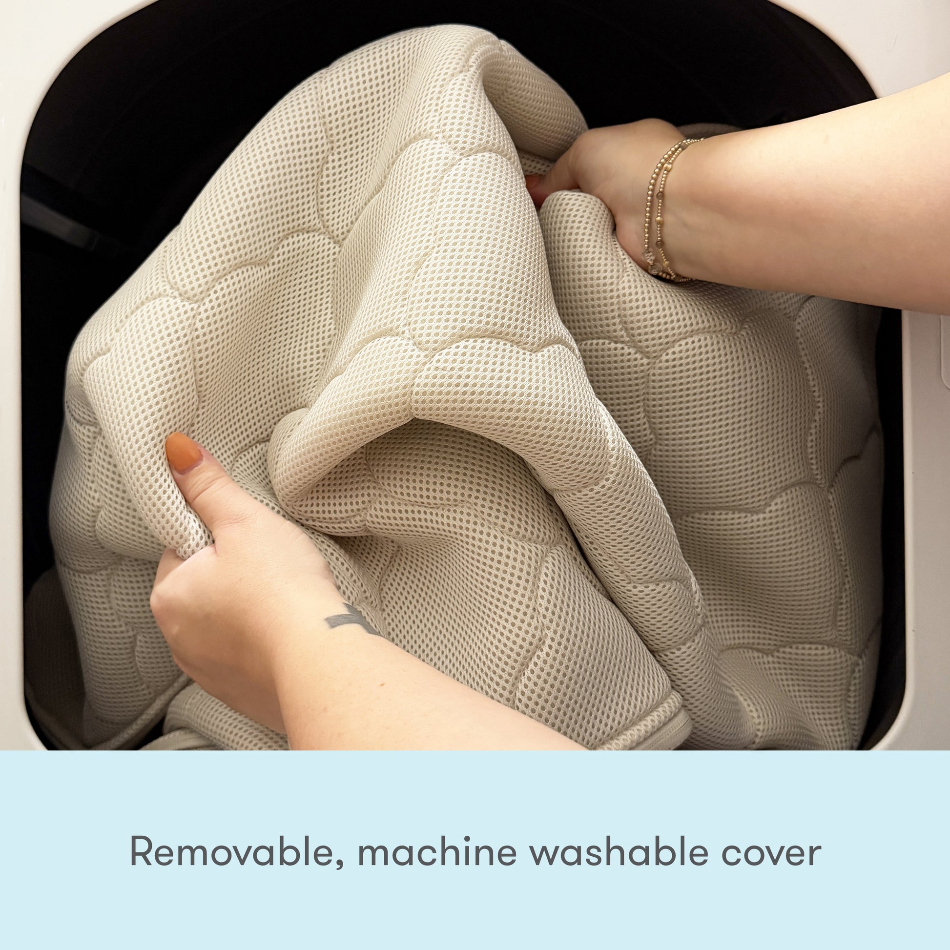 Close-up of hands placing a Newton Baby Waterproof Crib Mattress cover into a washing machine. Text below reads: "Removable, machine washable cover.