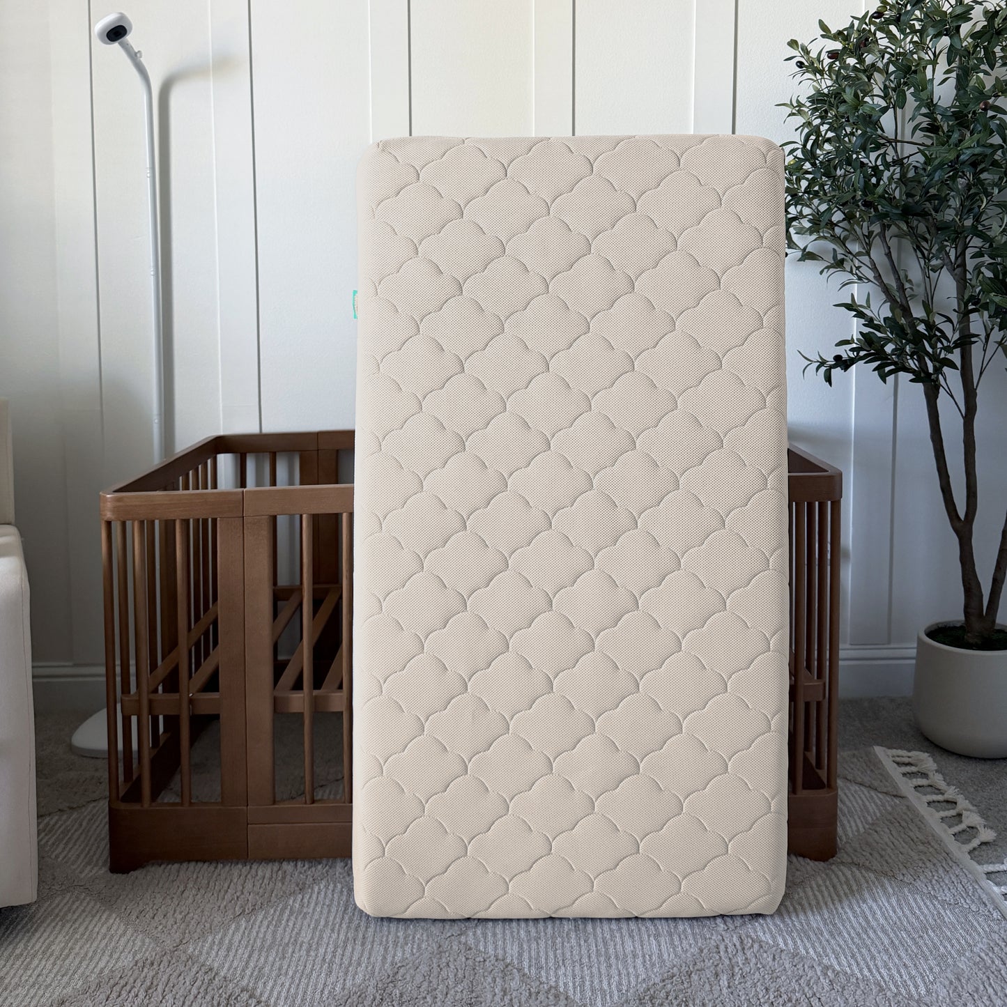 The Newton Baby Waterproof Crib Mattress, featuring a quilted beige design, stands upright in front of a wooden crib with a floor lamp and potted plant nearby, all set on a patterned rug.