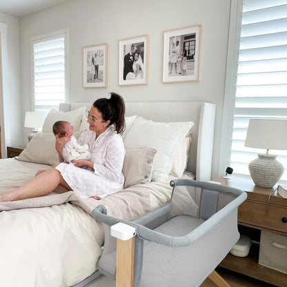 A smiling woman in pajamas gently interacts with a baby next to a gray bedside bassinet in a cozy bedroom setting.