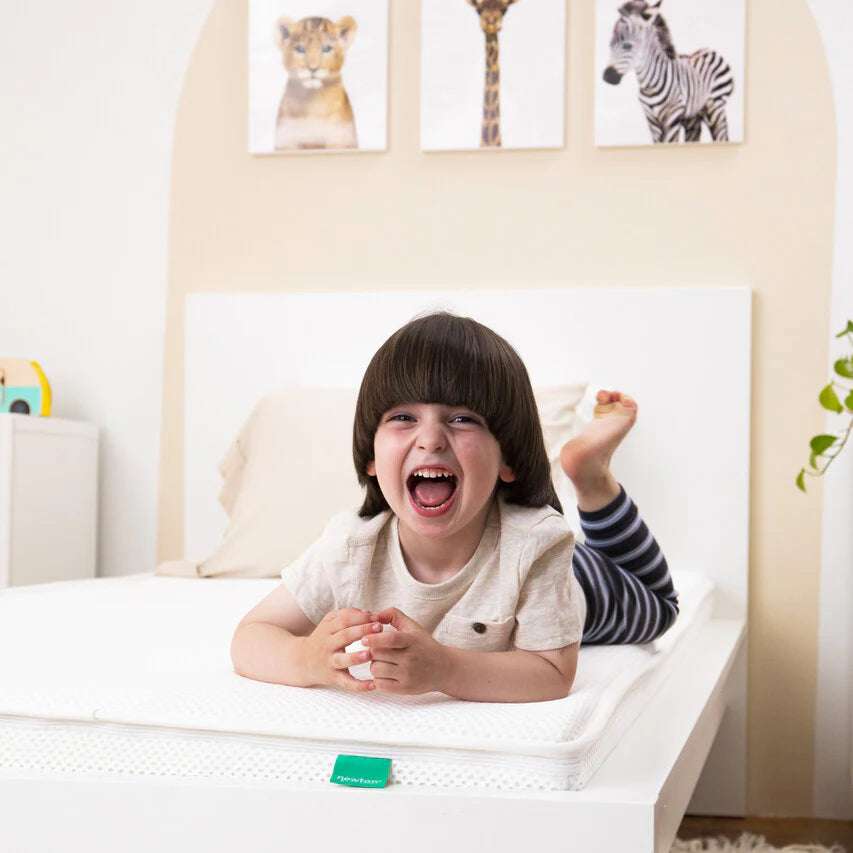 A young child with dark hair and striped pants lies on a white bed, smiling and laughing. Behind the child, animal art prints featuring a lion cub, giraffe, and zebra hang above the bed.