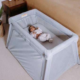 A baby in striped pajamas lies in a grey portable crib next to a bed with tan bedding and pillows, near a wooden nightstand and a plant on a light carpeted floor.