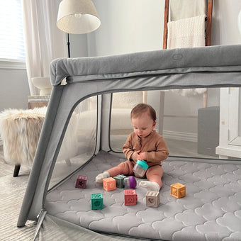 A baby sits contentedly inside a gray fabric-and-mesh playpen, dressed in a soft orange-brown onesie. Colorful silicone blocks and a light teal sippy cup dot the gray quilted mat at their feet. The background reveals a serene space: white curtains frame the scene, while a beige floor lamp and wooden ladder shelf (draped with neutral throws) complete the look. A cream faux fur rug peeks into the corner, adding warmth to the airy nursery.