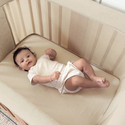 A baby rests on their back in a beige crib with Newton Baby Nest Bassinet Extra Sheets, wearing a light onesie and raising their left arm, gazing calmly toward the camera.