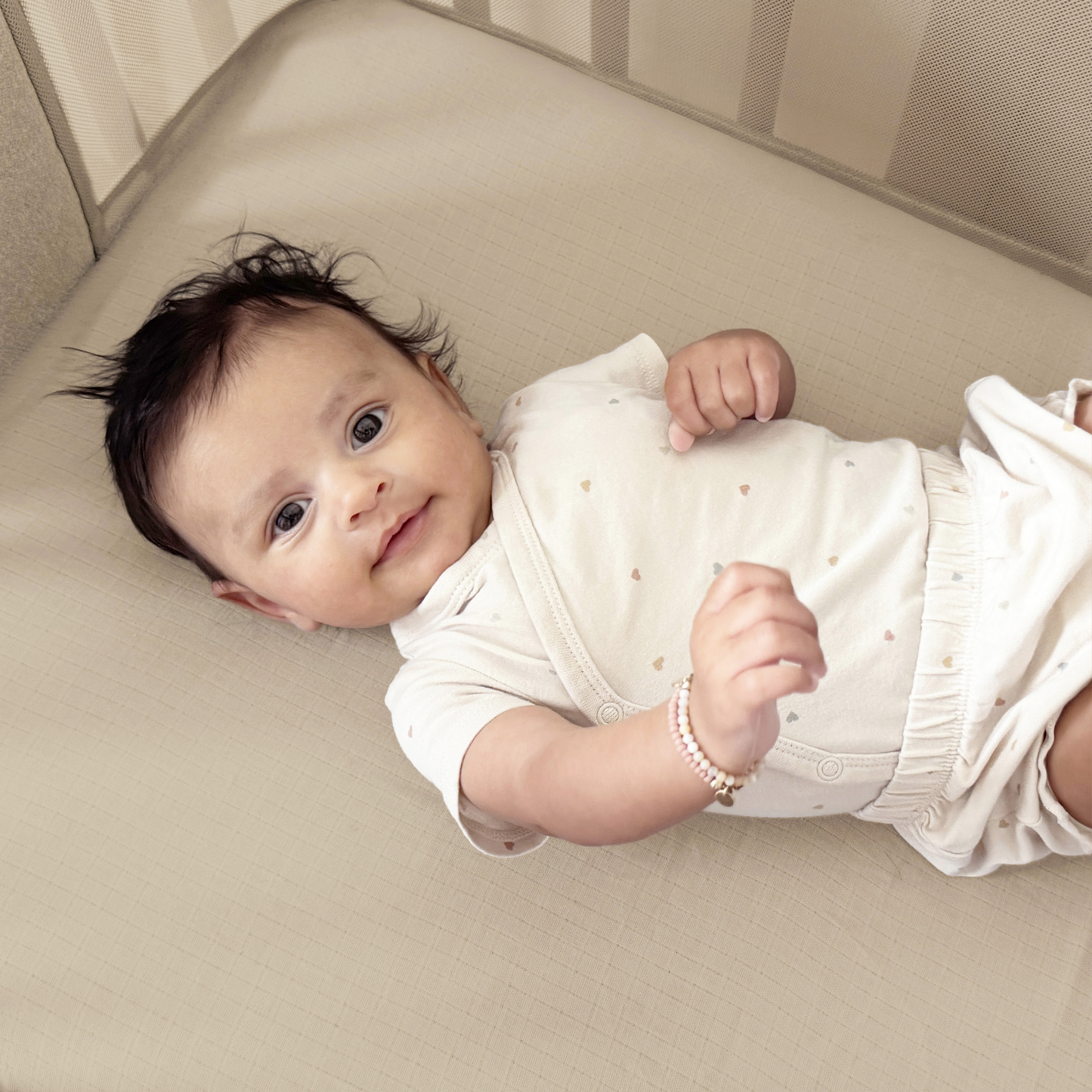 A baby with dark hair lies on its back, wearing a light onesie with small hearts and resting comfortably on Newton Baby Nest Bassinet Extra Sheets—soft, breathable crib sheets designed for comfort and safety. 999-8441-SAN