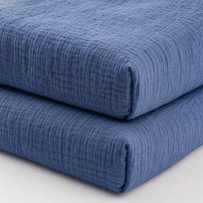 Two neatly folded Newton Baby Breathable, Organic Cotton Sheets (2-pack) in blue create a cozy, inviting look when stacked together on a light surface.