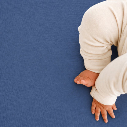 A baby in a cream-colored outfit sits on Newton Baby Breathable, Organic Cotton Sheets (2-pack) in soft blue, with only their legs, feet, and one hand visible.