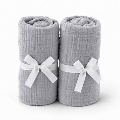 Two light gray towels, rolled and tied with white ribbons, stand side by side—an elegant pairing for Newton Baby's Breathable, Organic Cotton Sheets (2-pack), perfect for adding comfort and style to any serene nursery.