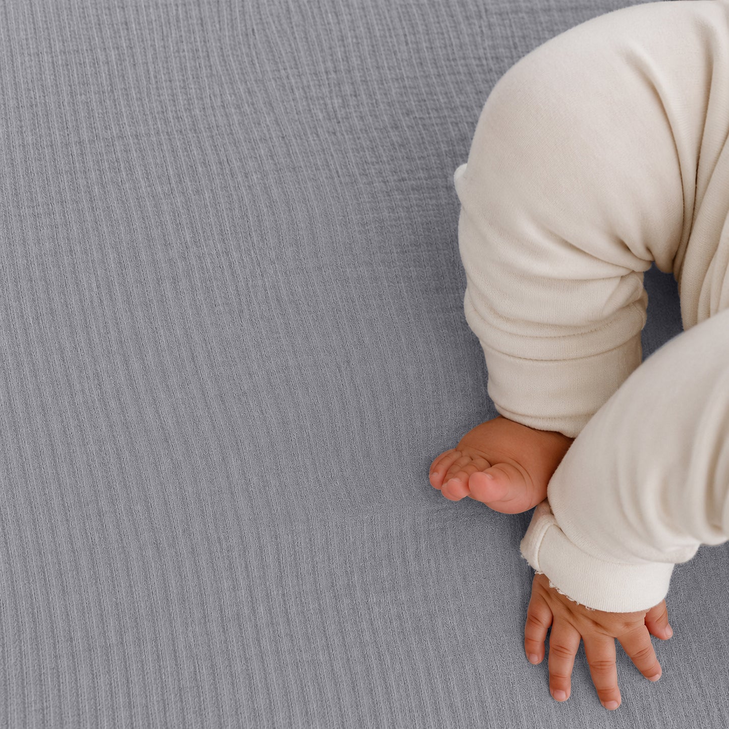 A baby in cream pajamas rests on Newton Baby Breathable, Organic Cotton Sheets (2-pack), with only the legs and one hand visible on the soft, gray bedding in the lower right of the image.