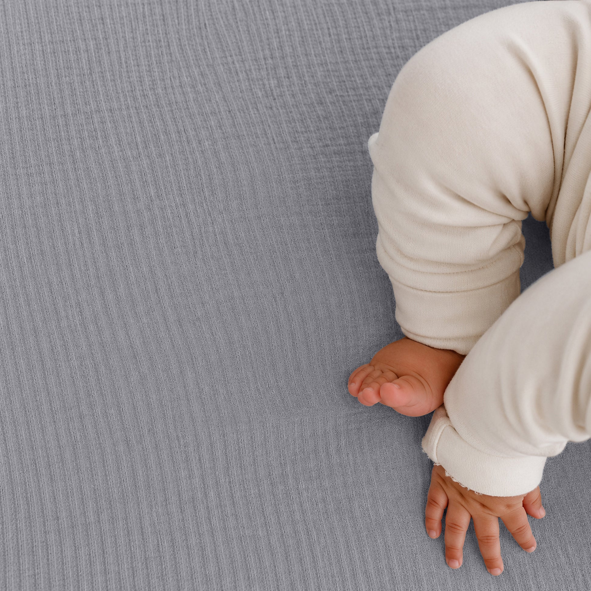A baby in cream pajamas rests on Newton Baby Breathable, Organic Cotton Sheets (2-pack), with only the legs and one hand visible on the soft, gray bedding in the lower right of the image.