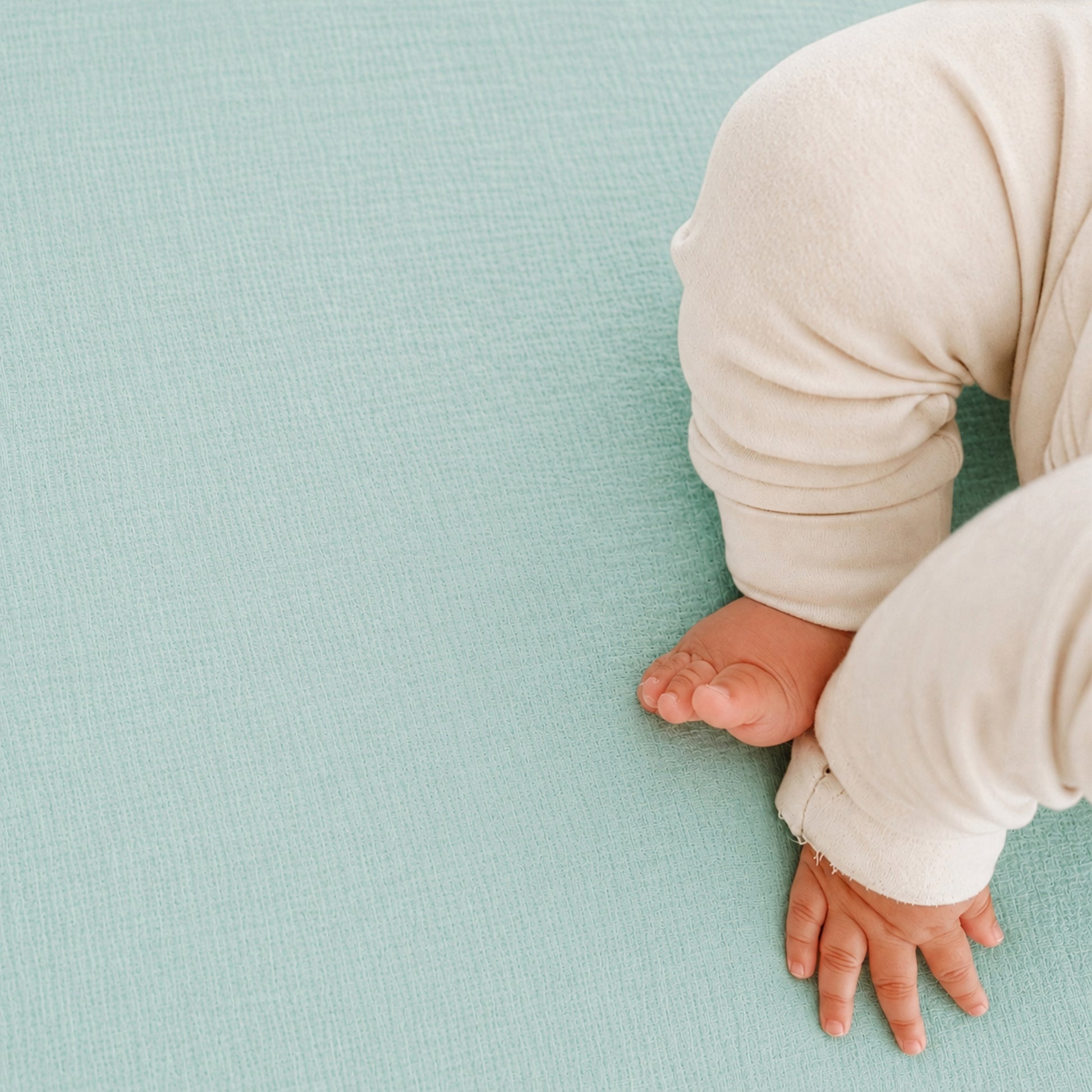 A close-up shows a baby's legs and hand resting on Newton Baby Breathable, Organic Cotton Sheets (2-pack) in light blue. The baby wears cream clothing, with only part of their body visible in the lower right corner.