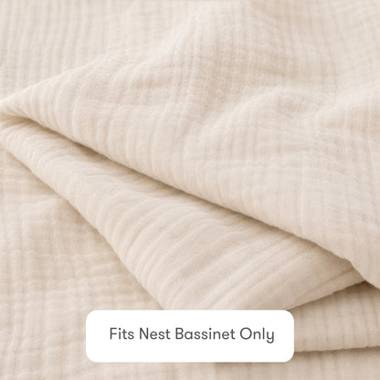 A close-up of a folded, cream-colored Newton Baby Nest Bassinet Extra Sheet made from textured cotton muslin. A label reads “Fits Nest Bassinet Only”—ideal for those wanting breathable sheets for their baby's crib.