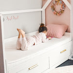 A toddler in a pink onesie lies on her stomach on a white daybed in a cozy, pink-themed room.