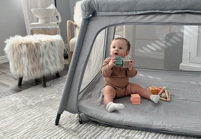 Baby in a brown onesie playing with toys inside a gray mesh playpen in a cozy room with a stylish chair and fur-covered stool.