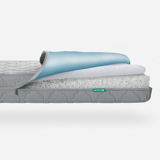 A cutaway of the Newton Baby Waterproof Crib Mattress shows its blue top cover, white middle layer, airy core, and a grey quilted washable outer cover with a small green label.