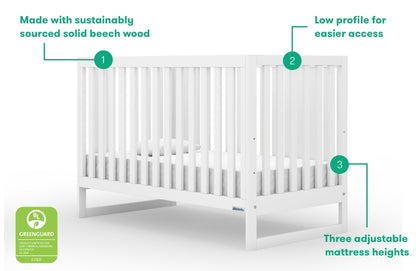 White solid beech wood crib with low profile for easier access, featuring three adjustable mattress heights and a GREENGUARD Gold certification label.
