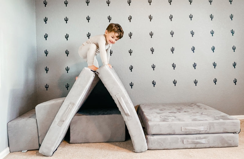 A young child in pajamas crouches on top of soft, gray foam play cushions arranged like a bridge or tunnel in a room with a cactus-patterned wall.