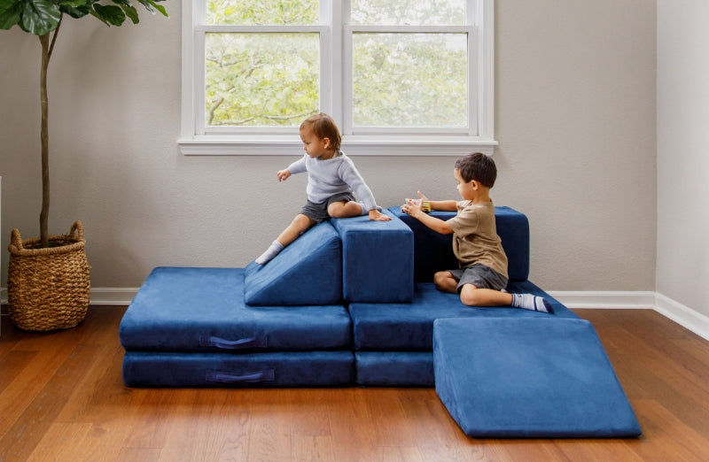 Two young children play on a modular blue foam play couch in a bright room with hardwood floors.