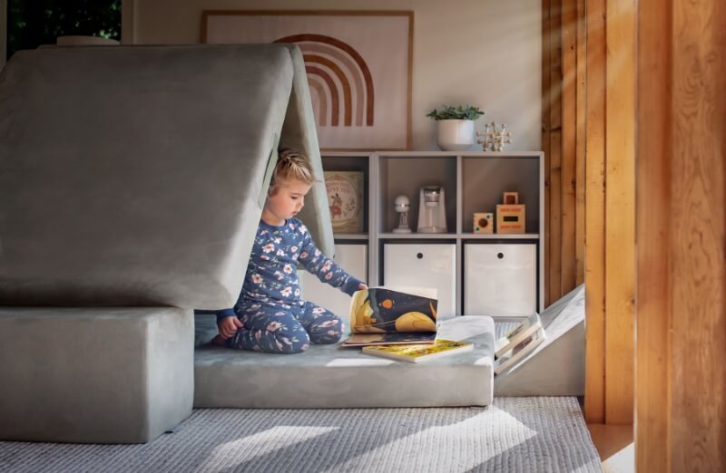 Toddler in pajamas reading a book in a cozy, sunlit room with stylish furniture and decor.