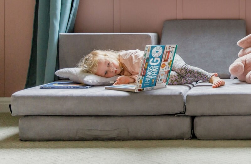 Toddler resting on a gray fold-out cushion with a book titled "Things That Go" covering her face in a cozy room.